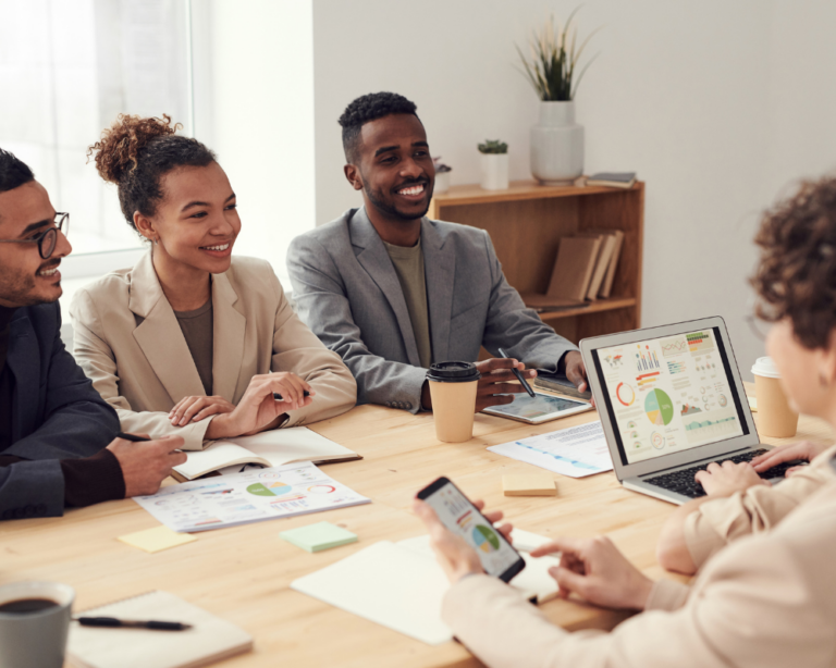 The image shows a diverse group of professionals in a business meeting, seated around a table in a well-lit office. Four individuals (two men and two women) are actively engaging in the discussion, smiling and looking towards a laptop displaying colorful graphs and data. Everyone is dressed in casual business attire. The atmosphere is collaborative and positive, reflecting a modern and dynamic work environment. To the right, there is a side table with plants, adding a touch of freshness and life to the workspace.